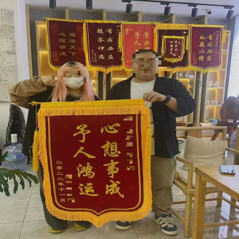 A smiling person holding a red and gold ceremonial banner with Chinese characters, representing a blessing for good luck and fortune, displayed in a bright indoor space.