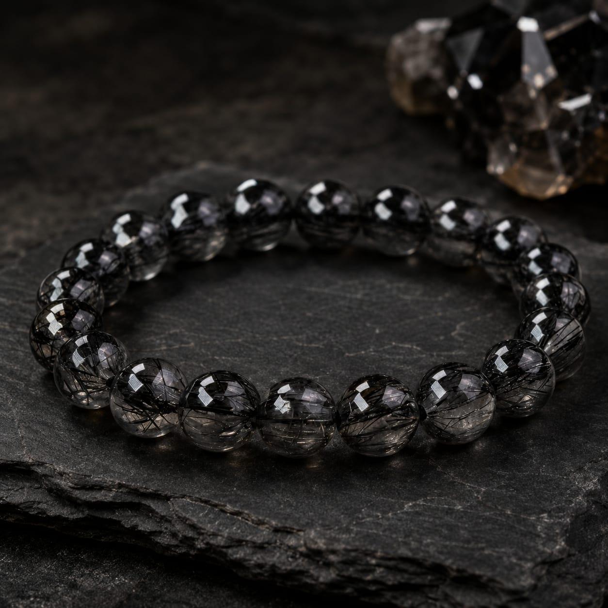 Natural Black Rutilated Quartz Bracelet displayed on a dark textured stone surface with black crystal accents, highlighting the smoky translucent beads and black needle-like inclusions.