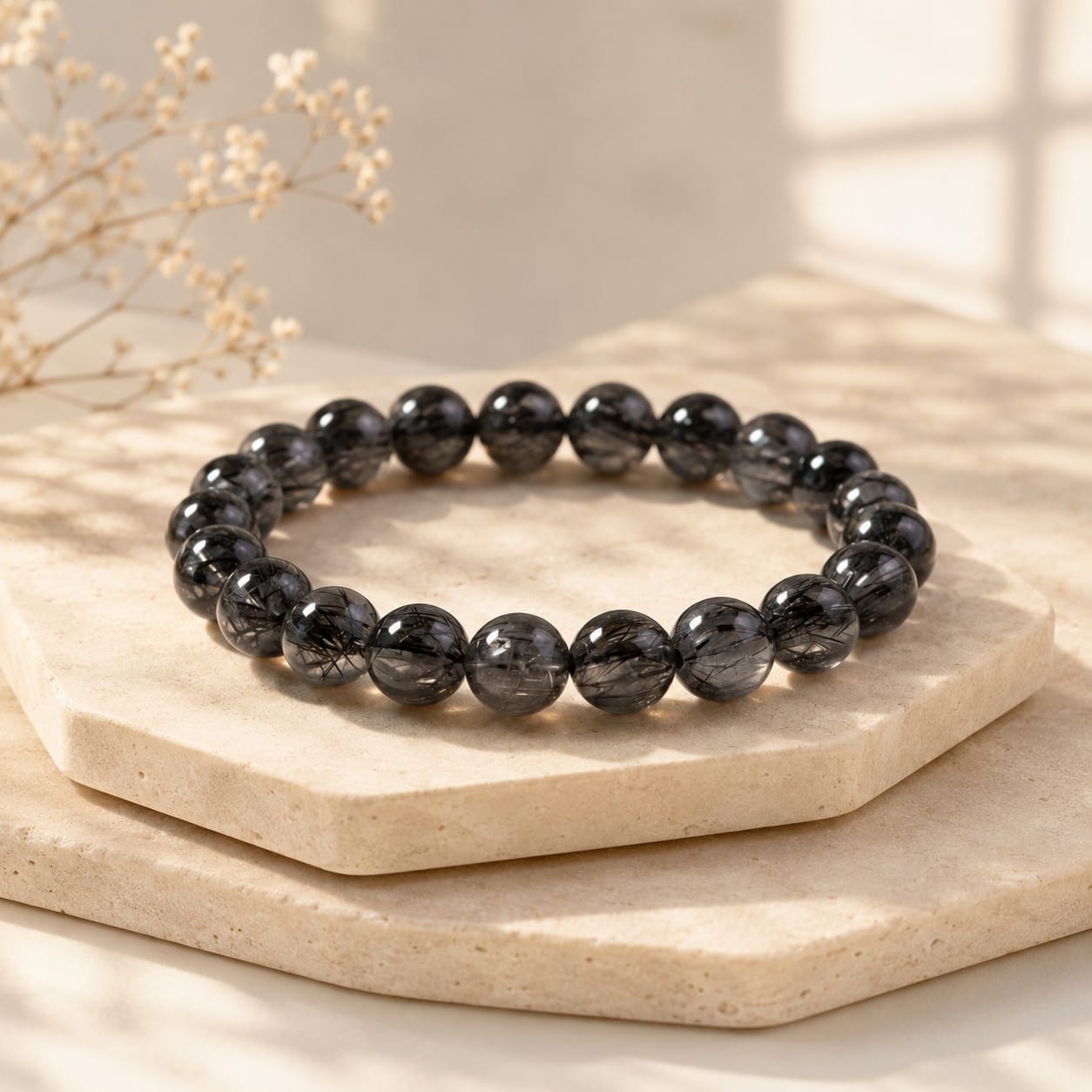 Natural Black Rutilated Quartz Bracelet displayed on layered light stone platforms in soft sunlight, emphasizing the bracelet’s refined shape and the dark inclusions inside each bead.