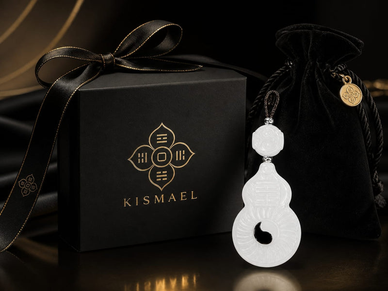 A luxurious black gift box beside a finely carved white Vermilion Bird charm pendant, placed next to a black velvet pouch. The box features the gold “KISMAEL” logo, with soft golden fabric in the background, highlighting an elegant and premium presentation.