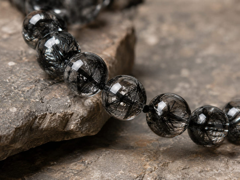 Close-up of a section of the Natural Black Rutilated Quartz Bracelet on a textured stone surface, highlighting the needle-like inclusions and deep smoky tones of the beads.