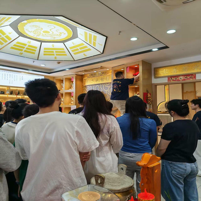 A group of people gathered inside a decorated room participating in a traditional festival or ceremony, some observing a ritual or presentation, with cultural decorations visible in the background.