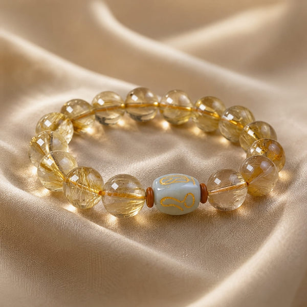 A detailed close-up of the Golden Rutilated Quartz Bracelet placed on a soft, cream-colored satin surface, highlighting the golden rutile strands and central San He Metal Formation bead.