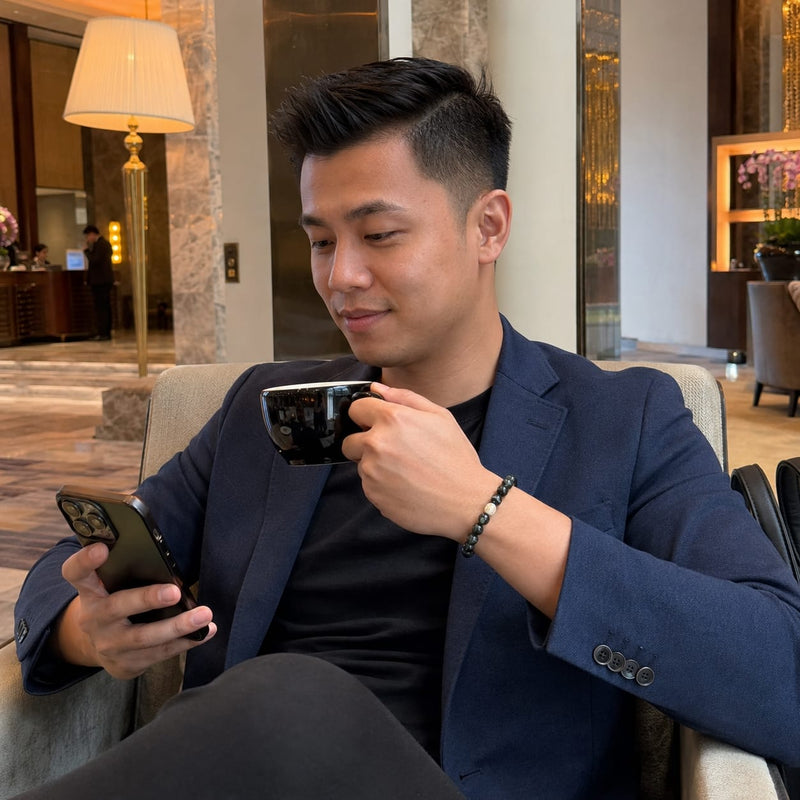 A man wearing a Green Phantom Quartz Bracelet while relaxing with coffee and checking his phone in a luxury lounge, symbolizing everyday abundance, calm confidence, and refined success.