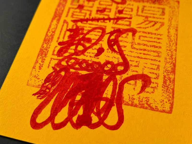 A macro view of a yellow hand-drawn Wealth Talisman with flowing red brush lines over a seal-style pattern. The image reveals the layered composition and traditional wealth symbolism, showcasing the refined detail and handcrafted Taoist-inspired design.