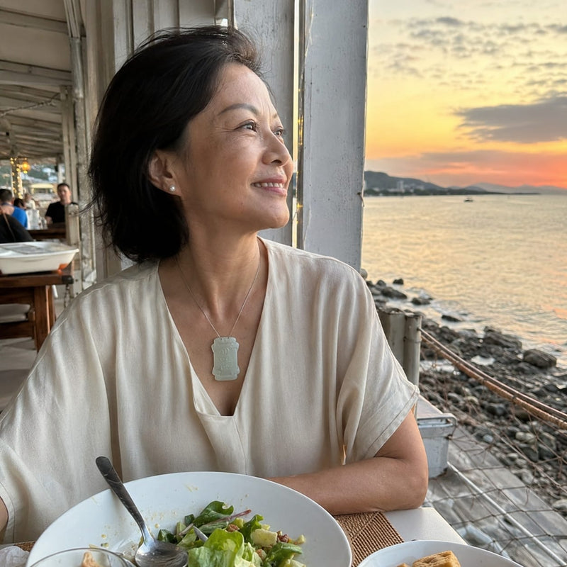 A mature overseas Chinese woman dining by the seaside at sunset, wearing the Moon Virtue Hetian jade pendant. The image reflects a comfortable, refined lifestyle and a sense of ease, balance, and quiet enjoyment.