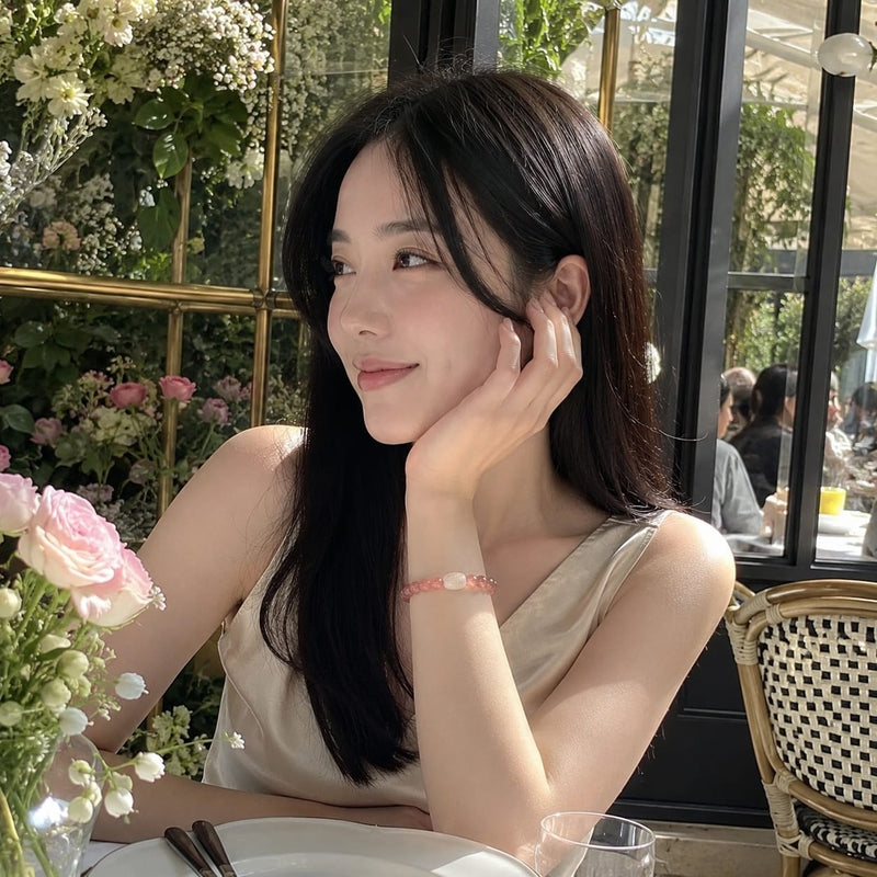 Young Asian woman sitting at a sunlit cafe table, wearing a Natural Rhodochrosite Bracelet with Hetian Jade talisman bead, looking contemplatively to the side amidst floral arrangements.
