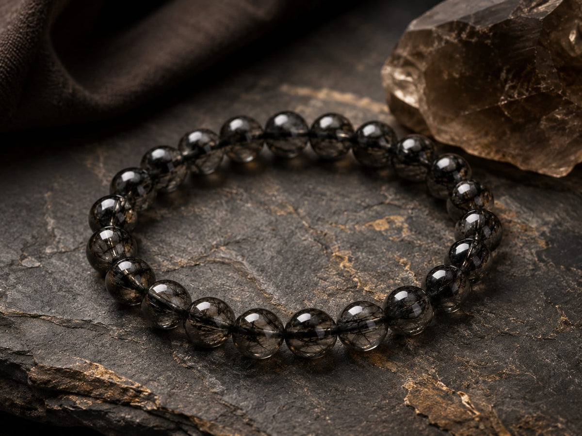 Natural Black Rutilated Quartz Bracelet placed on a dark textured stone surface, highlighting the smoky beads with black needle-like inclusions, symbolizing resilience, focus, and grounded energy.