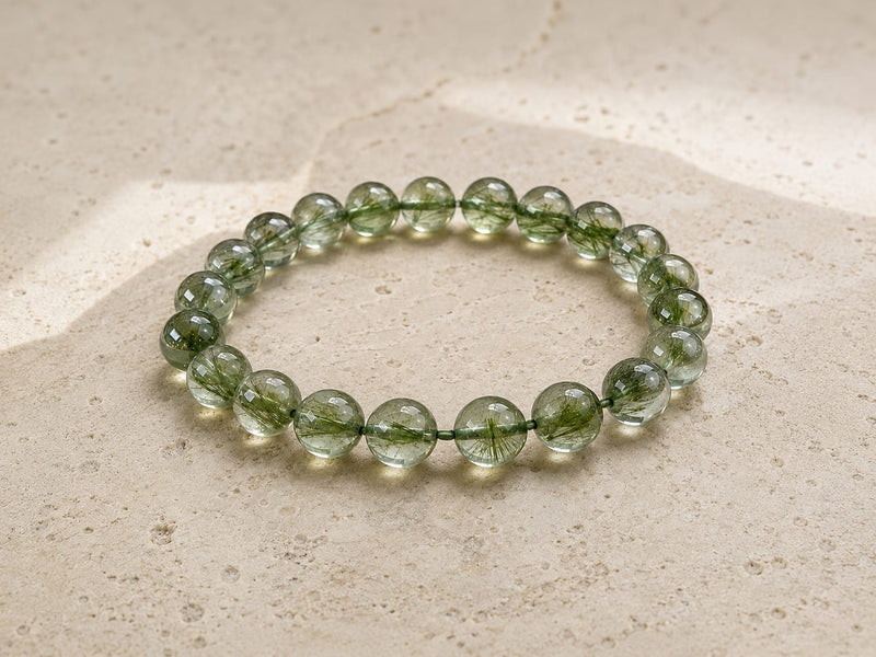 Natural green rutilated quartz bracelet displayed on a light stone surface, showing translucent green beads with delicate internal inclusions.