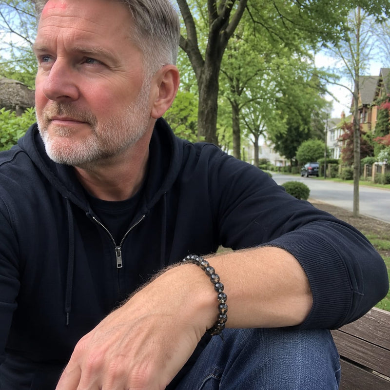 Middle-aged Caucasian man wearing a black rutilated quartz bracelet while sitting outside on a bench, reflecting on his surroundings, symbolizing balance and mindfulness.