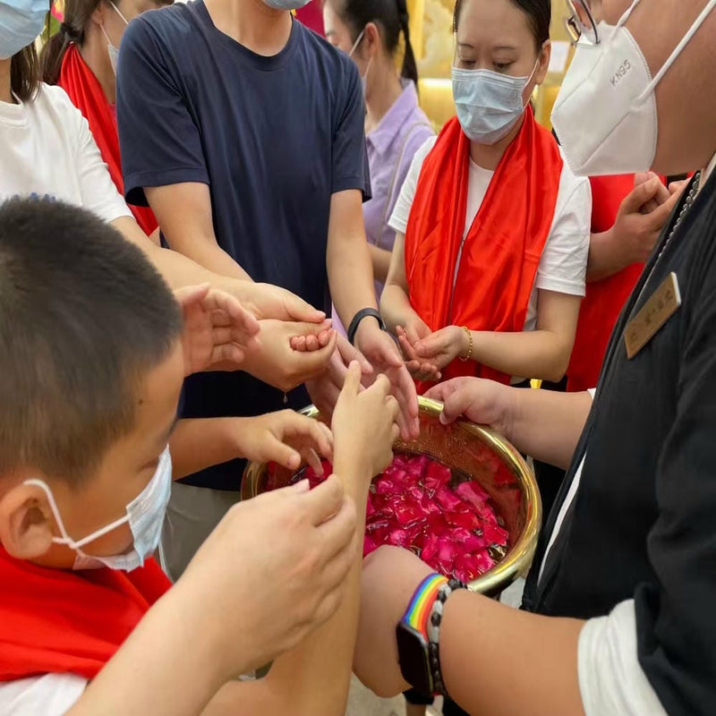 People performing a purification ritual by dipping hands into a bowl with petals under practitioner supervision.