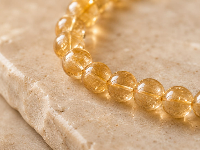 Close-up of polished citrine beads on a beige stone surface, catching the light and showing translucent golden tones with internal crystal textures.