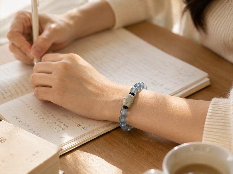 Close-up of a student’s hands wearing the Aquamarine Bracelet while reading and taking notes, with a cup of coffee and notebook nearby, capturing a calm, focused study moment.