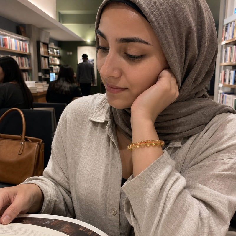 Malay woman wearing a Yellow Citrine Abundance Bracelet on her wrist while reading a book in a cozy indoor café. The golden beads are visible, reflecting warm light and adding subtle elegance.